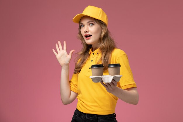 Vista frontal joven mensajero en uniforme amarillo sosteniendo tazas de café de plástico marrón con pose sobre fondo rosa trabajo uniforme entrega color trabajador de servicio