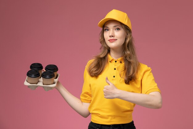 Vista frontal joven mensajero en uniforme amarillo sosteniendo tazas de café sobre fondo rosa oscuro trabajador de servicio de entrega uniforme