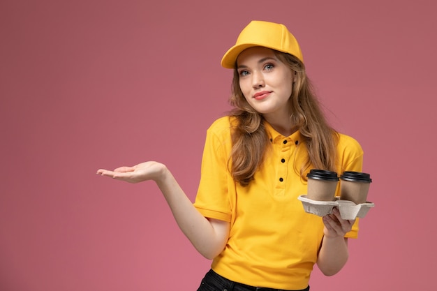 Vista frontal joven mensajero en uniforme amarillo sosteniendo tazas de café en el trabajador de servicio de entrega uniforme de escritorio rosa oscuro