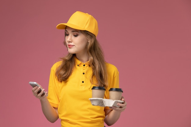 Vista frontal joven mensajero en uniforme amarillo sosteniendo tazas de café usando su teléfono en el escritorio de color rosa oscuro uniforme trabajador de servicio de trabajo de entrega