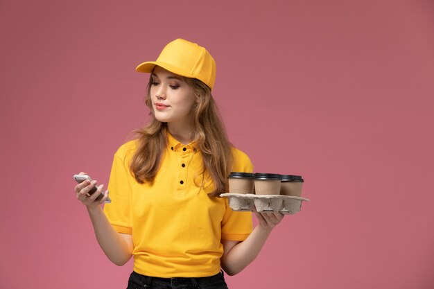 Vista frontal joven mensajero en uniforme amarillo sosteniendo tazas de café y usando su teléfono en la trabajadora del servicio de entrega uniforme de escritorio rosa oscuro