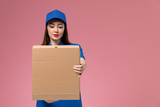 Vista frontal joven mensajero en uniforme azul y capa con apertura de caja de entrega de alimentos en la pared rosa claro