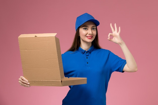 Vista frontal joven mensajero en uniforme azul y capa con apertura de caja de entrega de alimentos en la pared rosa claro