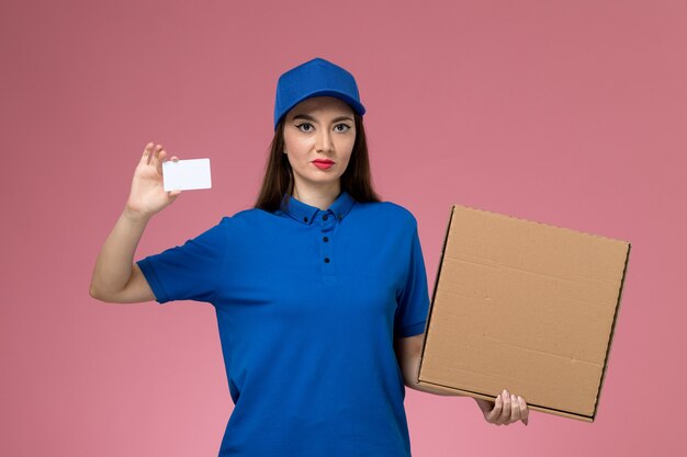 Vista frontal joven mensajero en uniforme azul y capa con caja de comida y tarjeta en la pared rosa