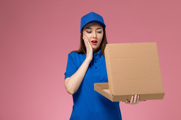 Vista frontal joven mensajero en uniforme azul y capa con caja de entrega de alimentos abriéndola en la pared rosa