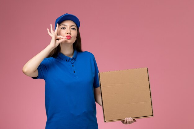 Vista frontal joven mensajero en uniforme azul y capa con caja de entrega de alimentos posando en la pared rosa