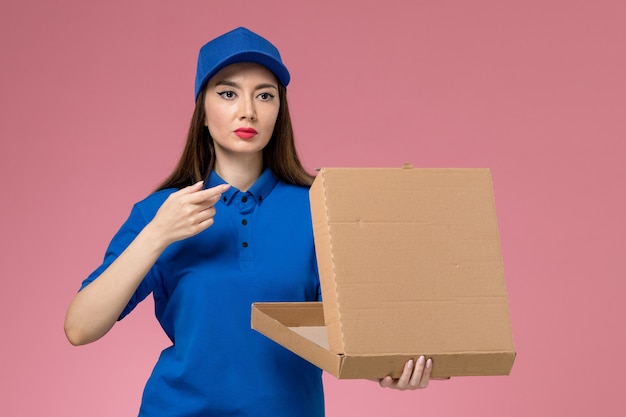 Vista frontal joven mensajero en uniforme azul y capa con caja de entrega de comida vacía en la pared rosa