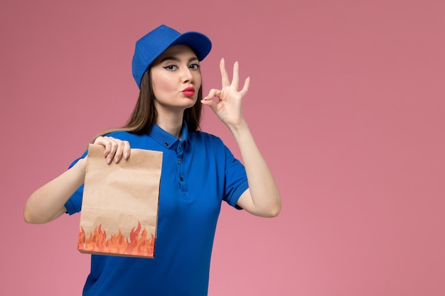 Vista frontal joven mensajero en uniforme azul y capa con paquete de comida de papel en la pared rosa