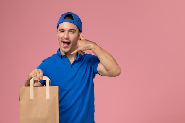 Vista frontal joven mensajero en uniforme azul y capa con paquete de entrega de papel en sus manos en la pared rosa