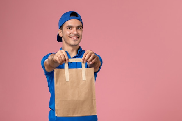 Vista frontal joven mensajero en uniforme azul y capa con paquete de entrega de papel en sus manos en la pared rosa