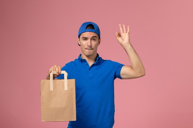 Vista frontal joven mensajero en uniforme azul y capa con paquete de entrega de papel en sus manos en la pared rosa