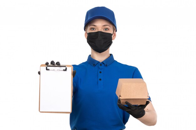 Una vista frontal joven mensajero en uniforme azul máscara negra y guantes con paquete de entrega de alimentos y bloc de notas