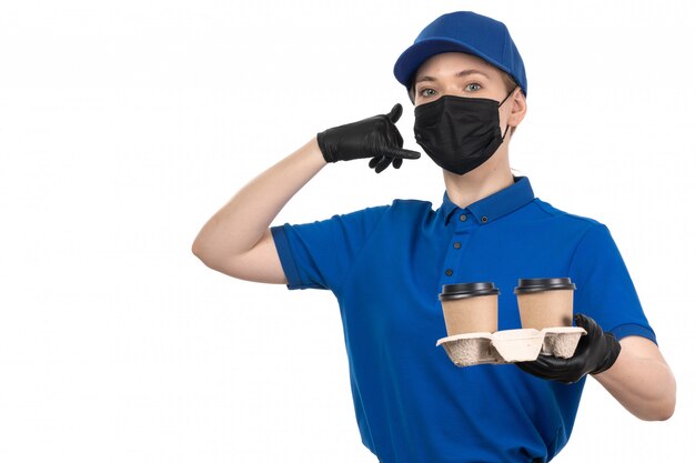 Una vista frontal joven mensajero en uniforme azul máscara negra y guantes sosteniendo tazas de café