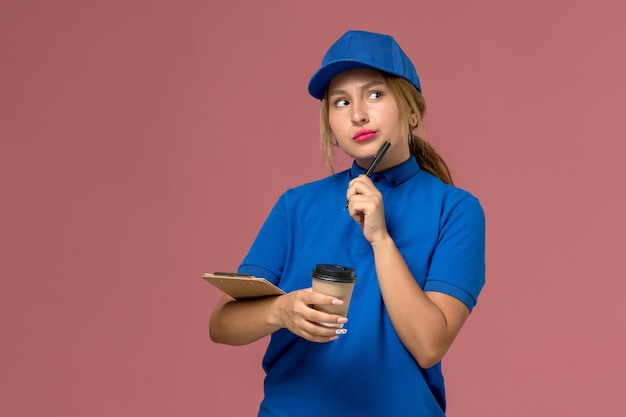 Vista frontal joven mensajero en uniforme azul posando sosteniendo una taza de café y un bloc de notas con expresión thiniking, mujer de entrega uniforme de servicio