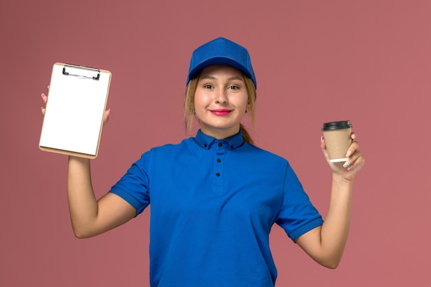 Vista frontal joven mensajero en uniforme azul posando sosteniendo una taza de café y bloc de notas, servicio de entrega uniforme trabajadora de trabajo