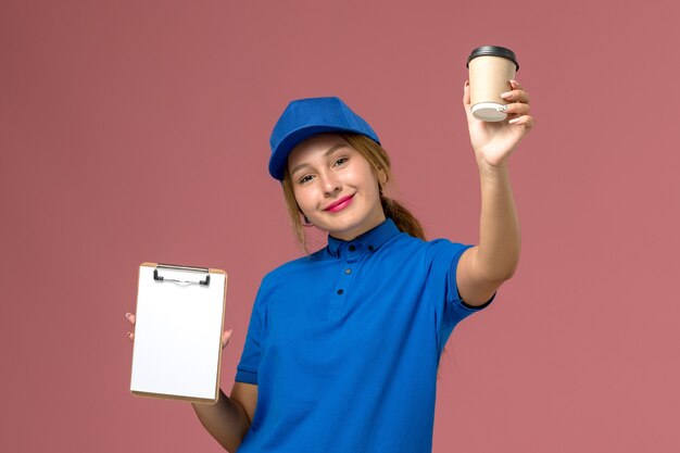 Vista frontal joven mensajero en uniforme azul posando sosteniendo una taza de café y un bloc de notas con una sonrisa, servicio uniforme mujer de entrega