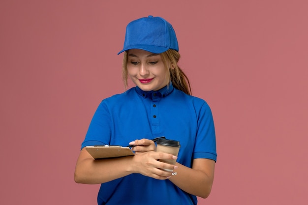 Vista frontal joven mensajero en uniforme azul posando sosteniendo una taza de café y bloc de notas, trabajadora de entrega uniforme de servicio