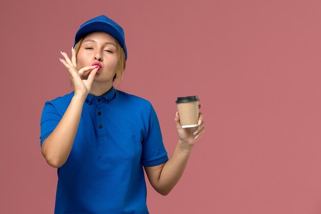 Vista frontal joven mensajero en uniforme azul posando sosteniendo una taza de café de entrega marrón en la pared rosa, entrega uniforme de trabajo de servicio