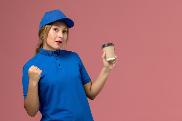 Vista frontal joven mensajero en uniforme azul posando sosteniendo una taza de café de entrega marrón en la pared rosa, mujer de entrega uniforme de trabajo de servicio