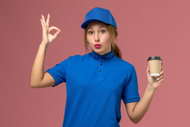 Vista frontal joven mensajero en uniforme azul posando sosteniendo una taza de café de entrega marrón en la pared rosa, mujer de entrega uniforme de trabajo de servicio