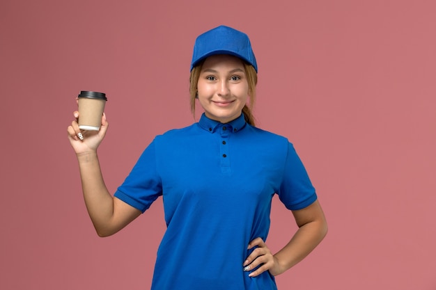 Vista frontal joven mensajero en uniforme azul posando y sosteniendo la taza de café de entrega en la pared rosa, mujer de entrega uniforme de trabajo de servicio