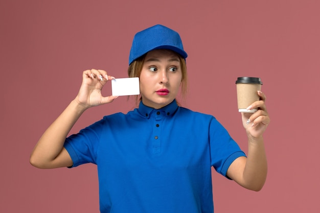 Vista frontal joven mensajero en uniforme azul posando sosteniendo una taza de café y tarjeta blanca, trabajador de trabajo de mujer de entrega uniforme de servicio