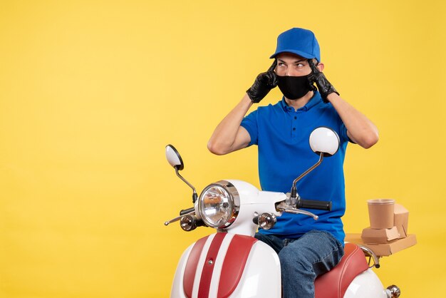 Vista frontal joven mensajero en uniforme azul sobre fondo amarillo covid- virus del trabajo pandémico entrega de trabajo en bicicleta