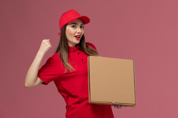 Vista frontal joven mensajero en uniforme rojo con caja de comida de entrega animando a la empresa de uniforme de entrega de servicio de fondo rosa