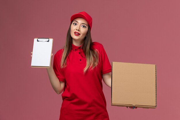 Vista frontal joven mensajero en uniforme rojo con caja de comida de entrega con bloc de notas sobre fondo rosa trabajador de empresa uniforme de servicio de entrega