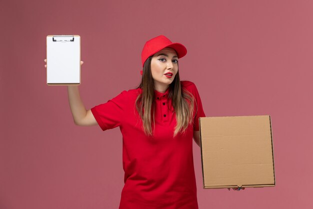 Vista frontal joven mensajero en uniforme rojo con caja de comida de entrega con bloc de notas en el trabajo de empresa uniforme de servicio de entrega de fondo rosa claro