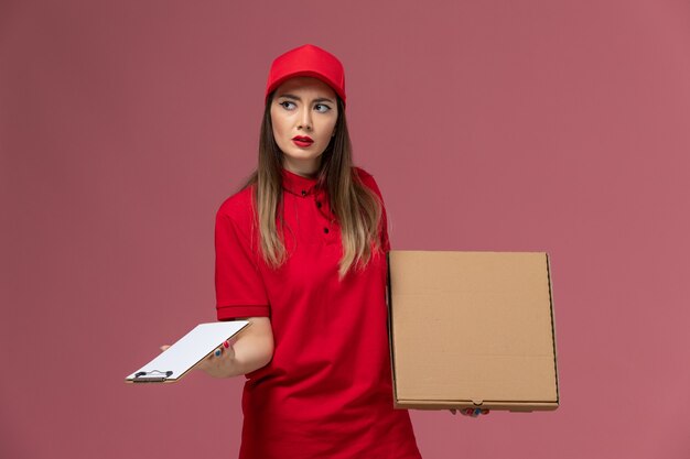 Vista frontal joven mensajero en uniforme rojo con caja de comida de entrega con bloc de notas en el trabajo de empresa uniforme de servicio de entrega de fondo rosa claro