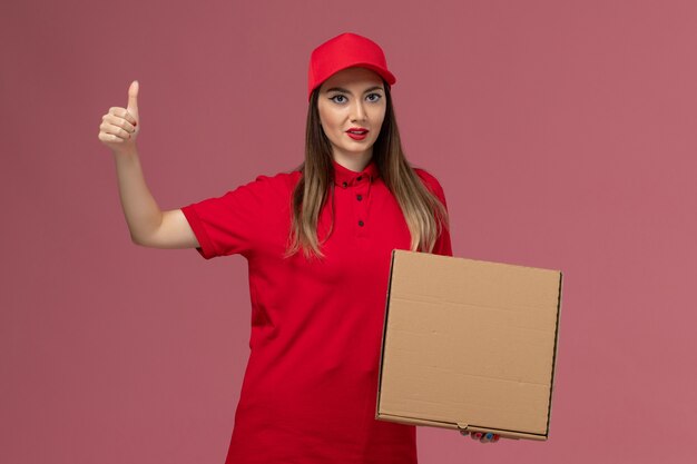 Vista frontal joven mensajero en uniforme rojo con caja de comida de entrega en la empresa de uniforme de entrega de servicio de fondo rosa