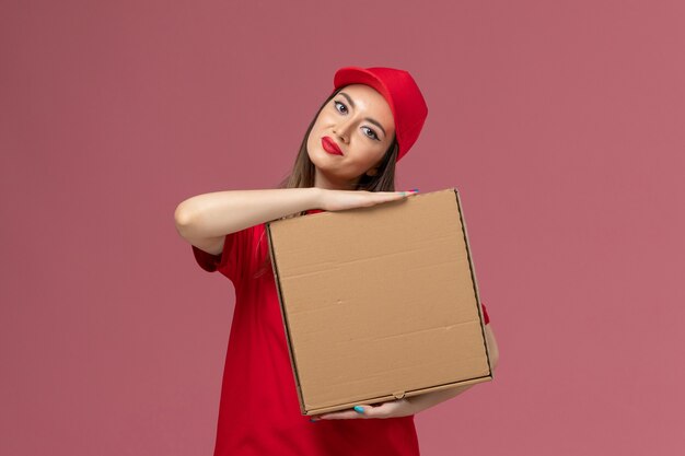 Vista frontal joven mensajero en uniforme rojo con caja de comida de entrega en la empresa de uniforme de entrega de servicio de piso rosa