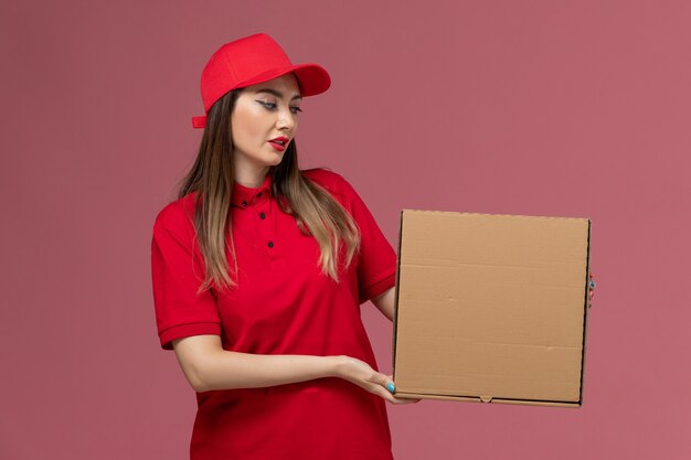 Vista frontal joven mensajero en uniforme rojo con caja de comida de entrega posando con él en la empresa de uniforme de entrega de servicio de fondo rosa