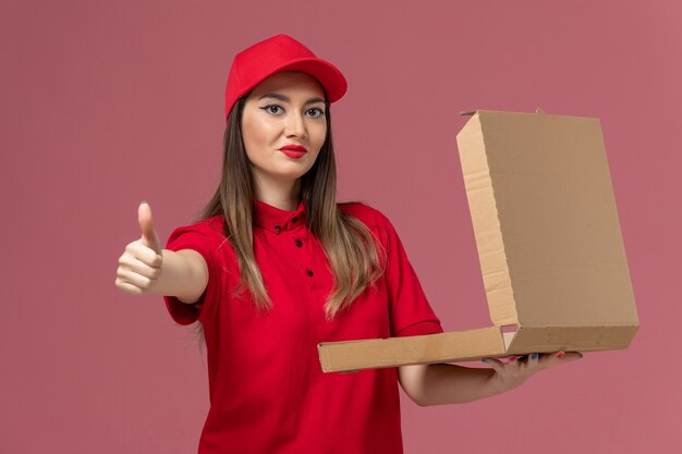 Vista frontal joven mensajero en uniforme rojo con caja de comida de entrega posando sobre fondo rosa claro servicio de entrega uniforme trabajador de la empresa