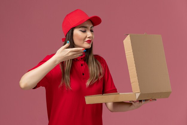 Vista frontal joven mensajero en uniforme rojo con caja de comida de entrega que huele a fondo rosa claro servicio de entrega uniforme trabajador empresa