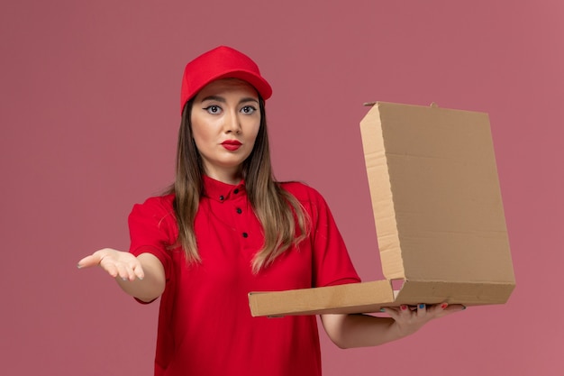 Vista frontal joven mensajero en uniforme rojo con caja de comida de entrega sobre fondo rosa claro servicio de entrega uniforme empresa de trabajadores