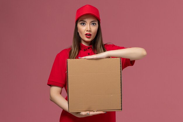 Vista frontal joven mensajero en uniforme rojo con caja de comida de entrega sobre fondo rosa empresa de uniforme de trabajo de entrega de servicio
