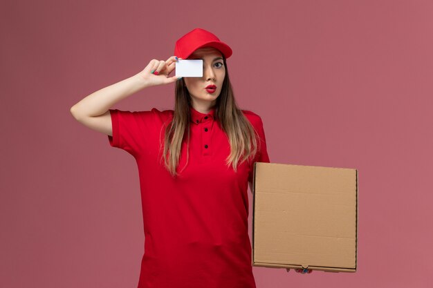 Vista frontal joven mensajero en uniforme rojo con caja de comida de entrega y tarjeta blanca sobre fondo rosa claro trabajo de empresa uniforme de servicio de entrega