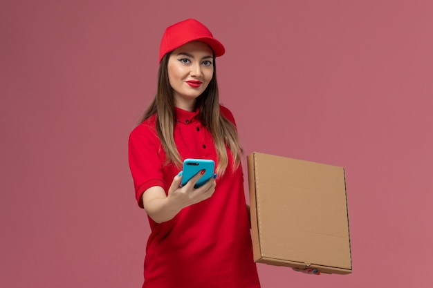 Vista frontal joven mensajero en uniforme rojo con caja de comida de entrega y teléfono en la empresa de trabajo uniforme de entrega de servicio de escritorio rosa