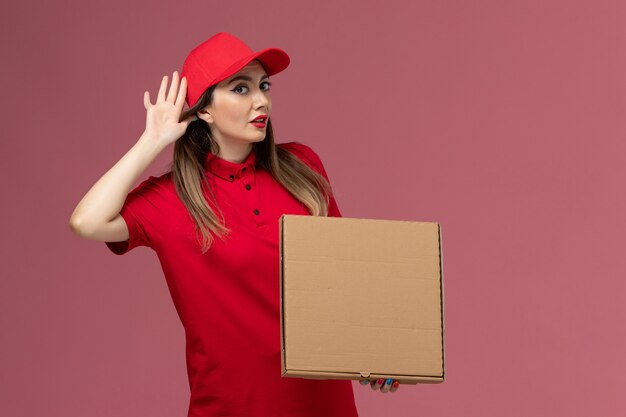 Vista frontal joven mensajero en uniforme rojo con caja de comida de entrega tratando de escuchar en la empresa de uniforme de entrega de servicio de fondo rosa