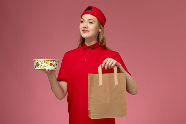 Vista frontal joven mensajero en uniforme rojo y capa con paquete de comida de entrega y tazón en pared rosa