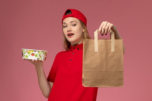 Vista frontal joven mensajero en uniforme rojo y capa con paquete de comida de entrega y tazón en servicio de escritorio rosa entrega uniforme de trabajo
