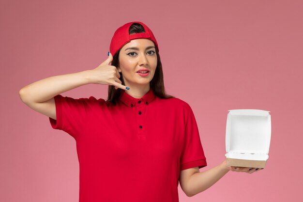 Vista frontal joven mensajero en uniforme rojo y capa con pequeño paquete de comida de entrega en sus manos en la pared rosa claro