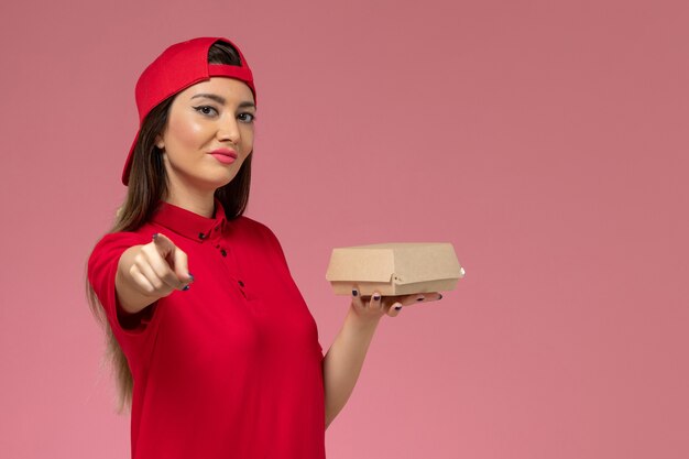 Vista frontal joven mensajero en uniforme rojo y capa con pequeño paquete de comida de entrega en sus manos en la pared rosa