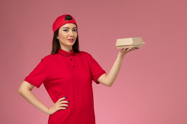 Vista frontal joven mensajero en uniforme rojo y capa con pequeño paquete de comida de entrega en sus manos en la pared rosa