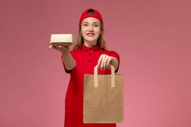 Foto gratuita vista frontal joven mensajero en uniforme rojo y capa sosteniendo paquetes de comida de entrega en la pared rosa