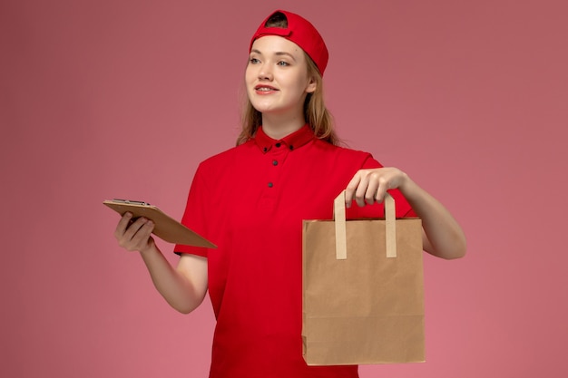 Vista frontal joven mensajero en uniforme rojo con paquete de comida de entrega y bloc de notas en la pared de color rosa claro