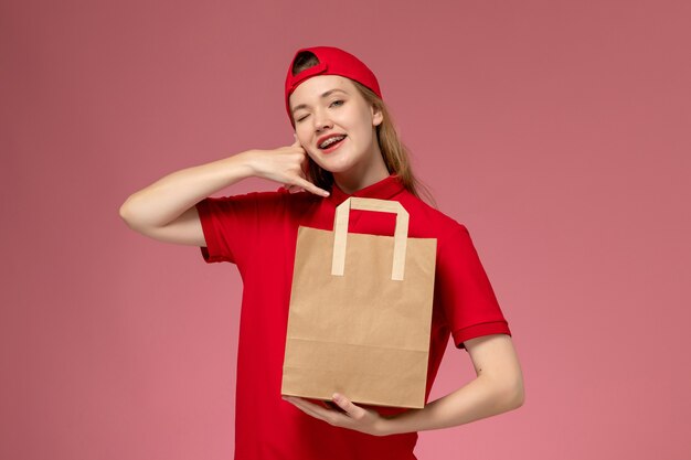 Vista frontal joven mensajero en uniforme rojo con paquete de comida de papel de entrega en la pared de color rosa claro