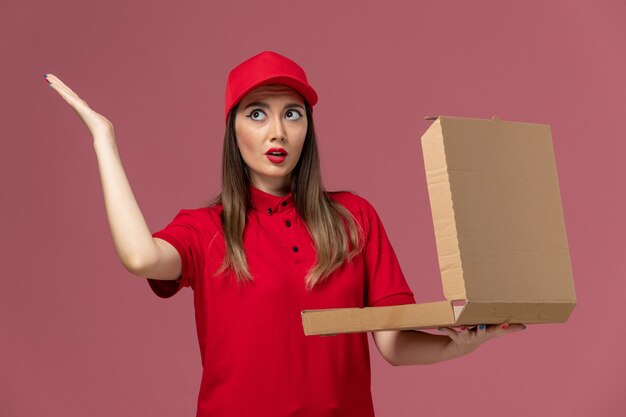 Vista frontal joven mensajero en uniforme rojo sosteniendo la caja de comida de entrega y abriéndola en la empresa de uniforme de trabajo de entrega de servicio de escritorio rosa claro